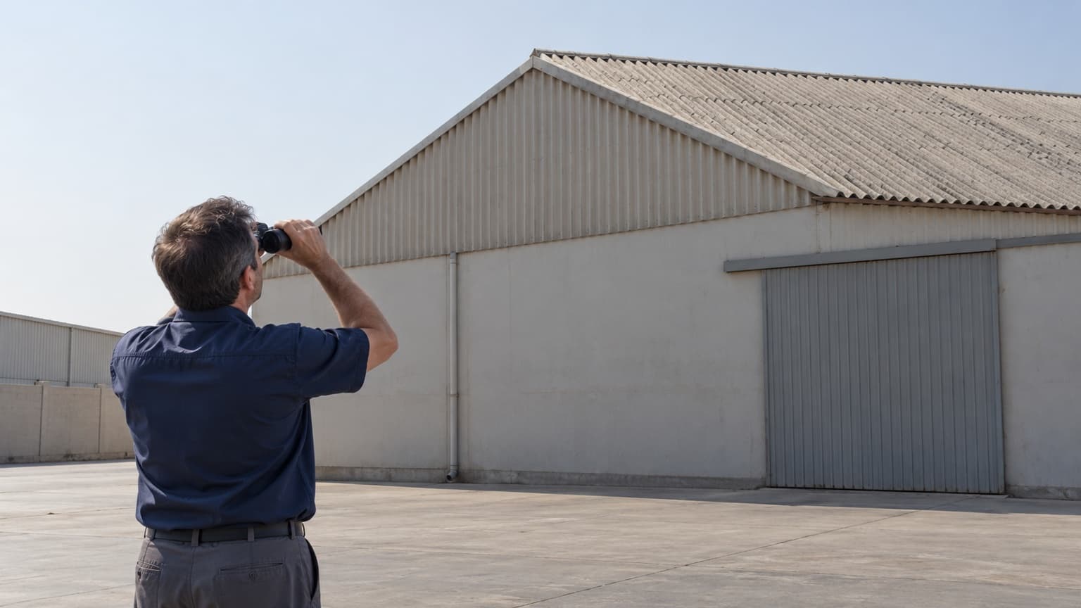 Illustrative facilities manager observing a corrugated warehouse roof from ground level with binoculars
